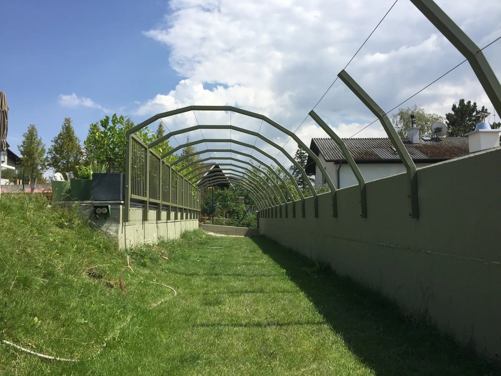 Gewölbte Metallbögen als Durchgang über einen Rasenweg, daneben ein grüner Zaun
